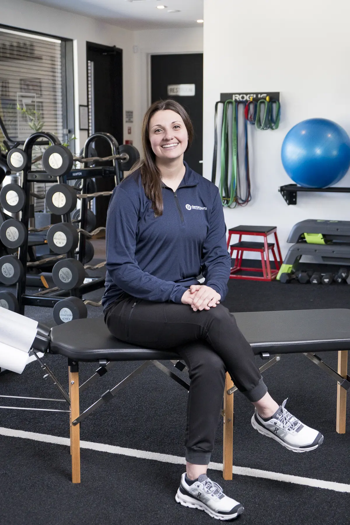 Dr. Abby Dante smiling, seated on a treatment table in a navy Dante Sports quarter-zip