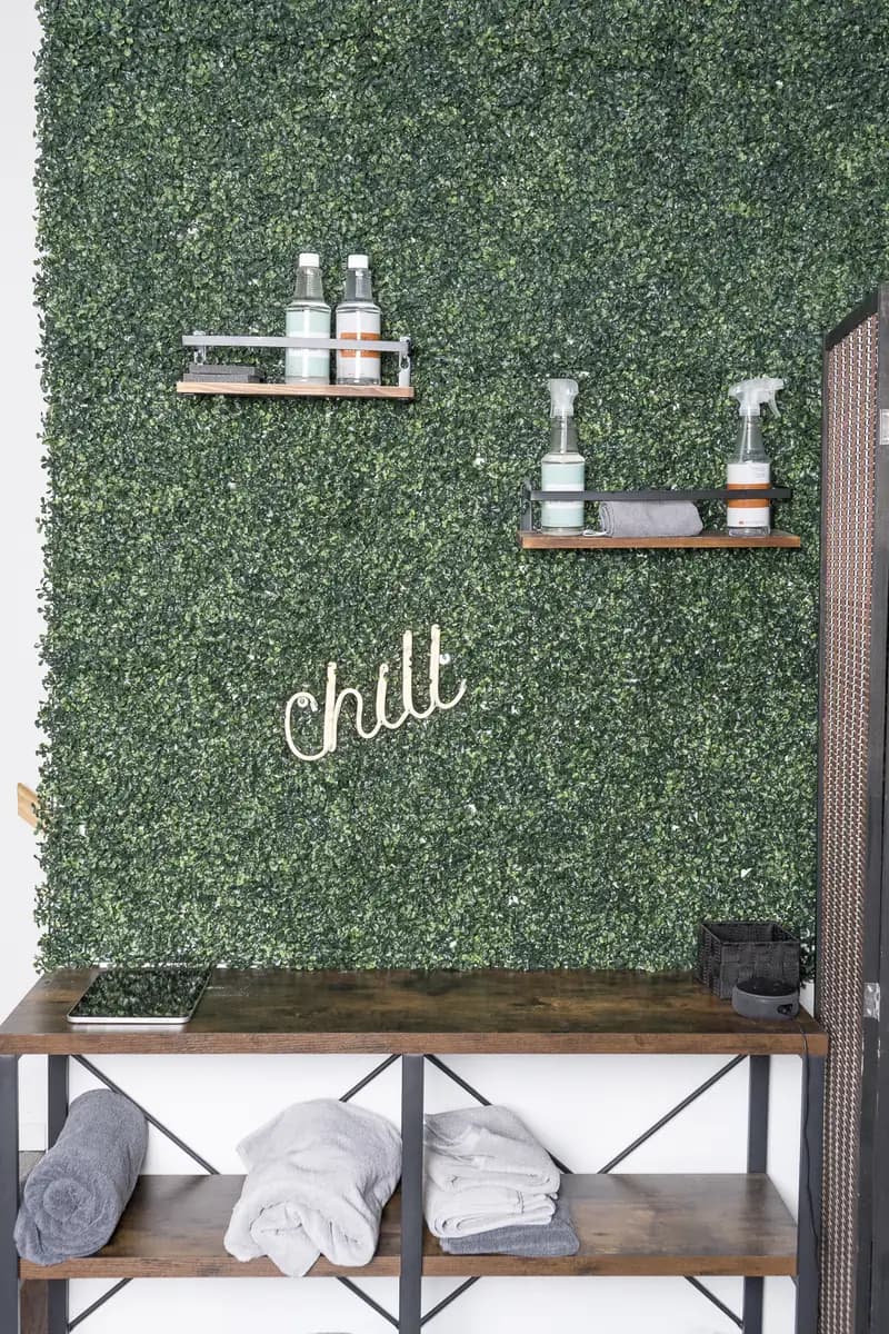 A boxwood wall with a "chill" sign and two small shelves of bottles, above a shelf of rolled towels